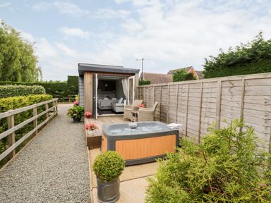 A small wooden cabin with an open door showing a bed inside a garden with a hot tub and outdoor chairs at Hydrangea Bretforton