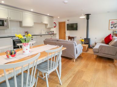 An open plan kitchen and living room with a wooden dining table and chairs a sofa a wood stove and kitchen cabinets at Shippon Barn in Yeovil