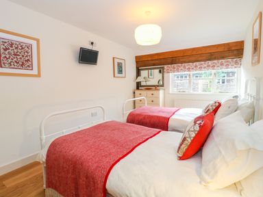 A bedroom with two single beds with red blankets and pillows a chest of drawers a wall-mounted television and a window with a patterned blind at Shippon Barn in Yeovil
