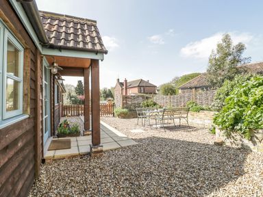 An outdoor patio area with gravel ground a wooden bench and metal chairs around a table and plants along a wooden house at Shippon Barn in Yeovil