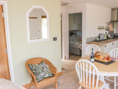 A kitchen and dining area with a wooden table and white chairs a wicker chair with a cushion and a bedroom visible through an open door at Black Cat Bach in Gilwern
