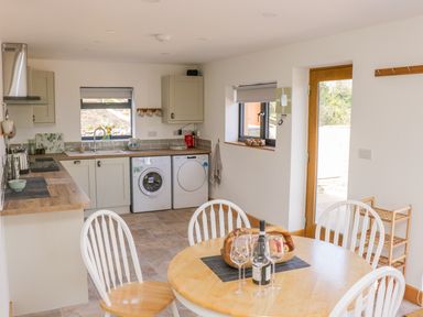 A kitchen and dining area with a round wooden table with wine glasses and a bottle and white chairs and a kitchen with appliances and a wooden door at Black Cat Bach in Gilwern