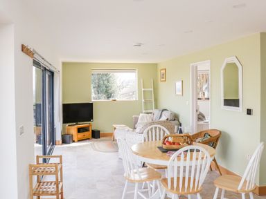A dining area with a wooden table and white chairs a living room with a sofa a television on a wooden stand and a wicker chair at Black Cat Bach in Gilwern