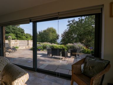 A room with a sofa and wicker chair looking out through large sliding glass doors onto a stone patio with outdoor furniture and garden at Black Cat Bach in Gilwern