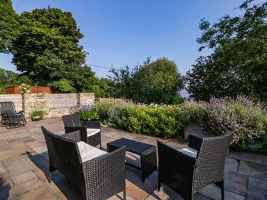 An outdoor patio with woven chairs and table surrounded by stone paving and greenery at Black Cat Bach in Gilwern