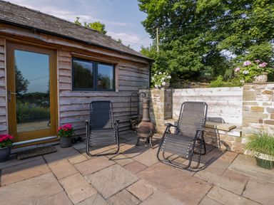 An outdoor patio with two black mesh chairs a small fireplace pots of flowers and a wooden door on a wooden building at Black Cat Bach in Gilwern