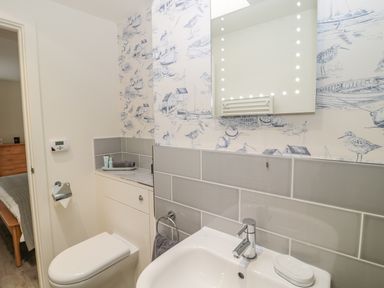 A bathroom with a toilet, a sink, a mirror with lights, and patterned wallpaper at Lambs Lodge in Bamburgh
