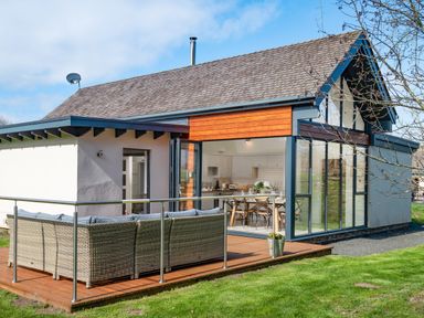 An outdoor patio with wicker seating and cushions next to a house with large glass windows and a wooden deck at Lambs Lodge in Bamburgh
