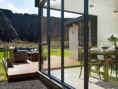 A dining area with a table set for meals visible through glass doors and an outdoor deck with seating furniture and a scenic rocky hill in the background at Lambs Lodge in Bamburgh