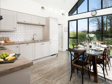 A kitchen with white cabinetry and a dining table set with plates and glasses by large windows at Lambs Lodge in Bamburgh