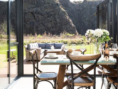 A dining table with wooden chairs set with plates and glasses near a patio with loungers and mountains in the background at Lambs Lodge in Bamburgh