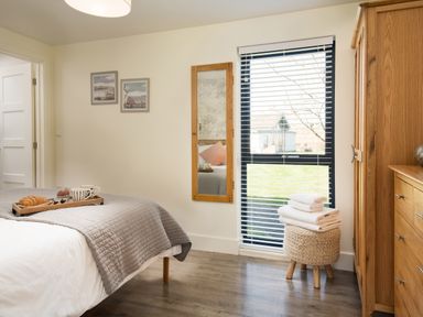 A bedroom with a bed covered with a gray quilt and breakfast tray a mirror on the wall window with blinds a woven stool with folded towels and wooden furniture at Lambs Lodge in Bamburgh