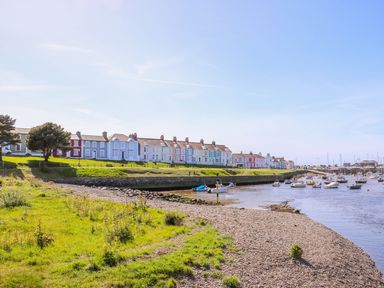 A shoreline with grass and pebbles leading to a body of water with boats moored near colorful houses at Bari in Aberaeron