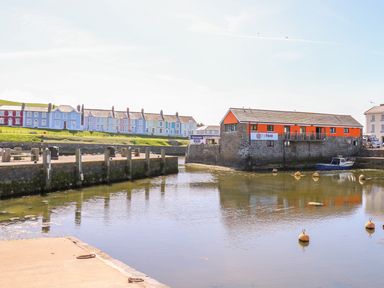 A harbor with a stone building with orange walls and boats on water with colorful houses in the background at Bari in Aberaeron