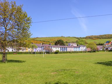 A row of colorful houses behind a large grassy field with trees and hills in the background at Bari in Aberaeron