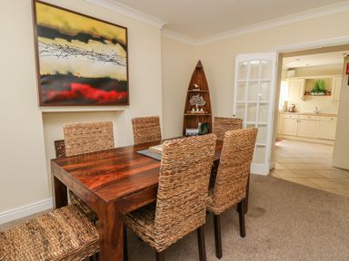A dining room with a wooden table and woven chairs a painting on the wall and a corner shelf near a door to a kitchen at Bari in Aberaeron