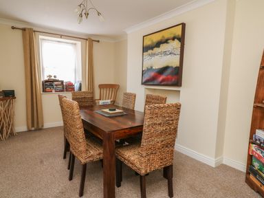 A dining room with a wooden table and woven chairs next to a window with curtains and a painting on the wall at Bari in Aberaeron