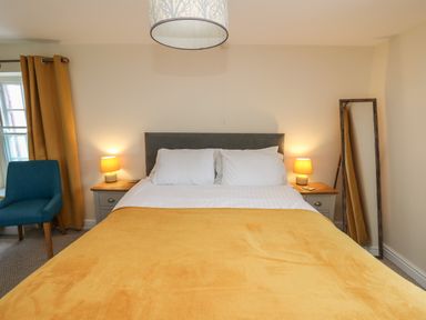 A bedroom with a bed with white pillows and a yellow blanket two bedside tables with lamps a blue chair yellow curtains and a standing mirror at Bari in Aberaeron