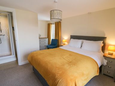 A bedroom with a double bed covered with a yellow blanket two bedside tables with lamps a blue chair a chest of drawers and an ensuite bathroom at Bari in Aberaeron