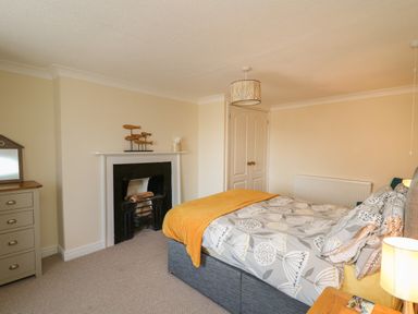 A bedroom with a double bed covered with a patterned duvet and orange throw a wooden dresser mirror a fireplace and a ceiling light at Bari in Aberaeron