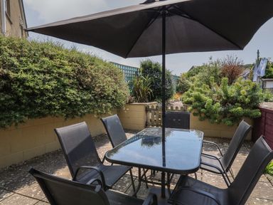An outdoor patio area with a glass table and six black chairs under a black umbrella surrounded by bushes and plants at Bari in Aberaeron