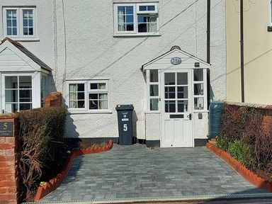 The front entrance of a white house with a small porch, paved driveway, a black bin, hedge borders, and a water butt at Meadow View Cottage in Lympstone