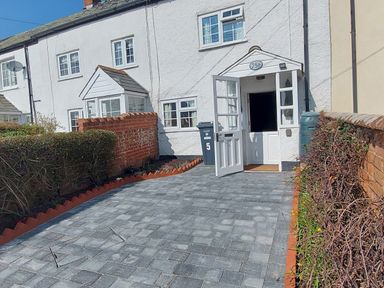 A white house with an open door a paved driveway and trimmed hedges at Meadow View Cottage in Lympstone