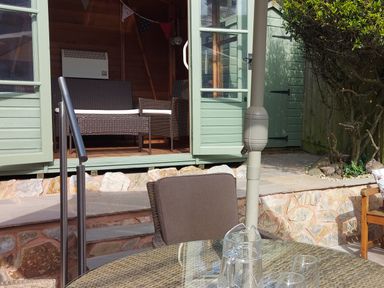An outdoor patio area with a glass table and chairs next to a green wooden garden shed with open doors showing wicker furniture inside at Meadow View Cottage in Lympstone