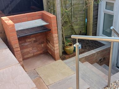 An outdoor brick barbecue next to stairs with metal railing and potted plant in a garden area at Meadow View Cottage in Lympstone