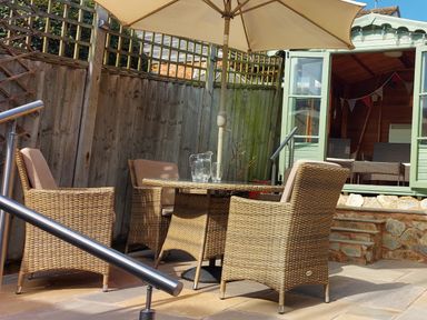 An outdoor patio with four wicker chairs around a glass table under a large umbrella at Meadow View Cottage in Lympstone