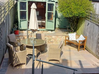 An outdoor patio with a round table and four chairs an umbrella a wooden bench with cushions and a garden shed at Meadow View Cottage in Lympstone