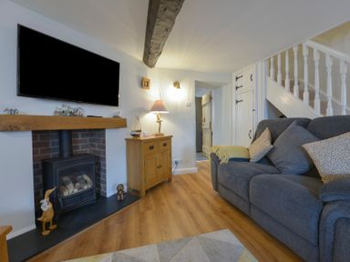 A living room with a gray sofa cushions and a wooden cabinet with lamp beside a wood stove with a television mounted above at Meadow View Cottage in Lympstone
