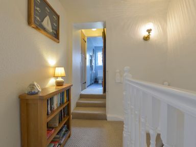 A hallway with carpeted stairs a wooden bookshelf with books a table lamp and wall light leading to a bathroom at Meadow View Cottage in Lympstone