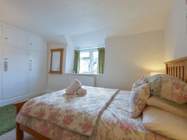 A bedroom with a floral bedspread on a wooden bed a window with green curtains built in white wardrobes and a wall mirror at Meadow View Cottage in Lympstone