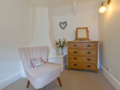 A corner with a pink upholstered chair with a cushion a wooden chest of drawers with a mirror on top and a vase of flowers on a shelf at Meadow View Cottage in Lympstone