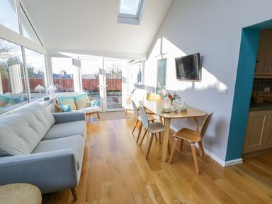 A room with a sofa, dining table with chairs, TV on the wall, and large windows letting in natural light at Clyd Fan in Harlech