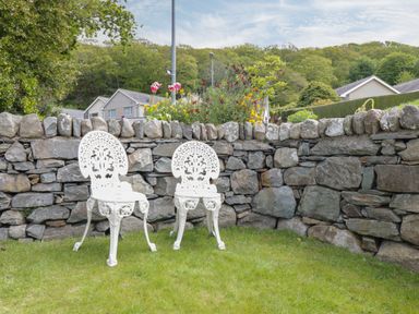 Two white metal chairs on grass in front of a stone wall with flowers and houses in background at Clyd Fan in Harlech