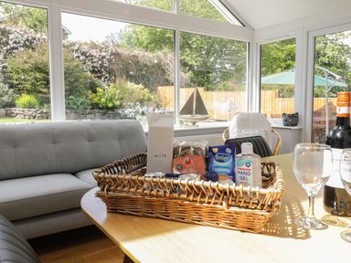 A dining table with a wicker basket containing milk chocolate bars tea and hand gel in a room with large windows and a gray sofa at Clyd Fan in Harlech