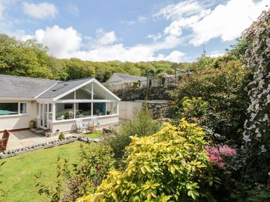 A backyard garden with green grass shrubs and flowers beside a house with a sunroom at Clyd Fan in Harlech
