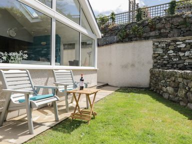 A small garden with two chairs and a wooden table with wine and glasses outside a house with large windows at Clyd Fan in Harlech