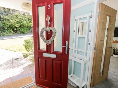 An open red front door with a heart-shaped wreath and a white coat rack with hooks next to an open wooden door leading into a room