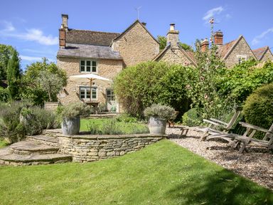 A stone house with a garden that has potted plants wooden chairs and an umbrella at The Old Post Office in Coln St Aldwyns