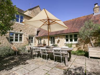 An outdoor patio with a wooden table chairs and a large umbrella attached to a stone house at The Old Post Office in Coln St Aldwyns