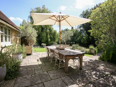 An outdoor patio with a wooden table and metal chairs under a large umbrella surrounded by plants and trees at The Old Post Office in Coln St Aldwyns