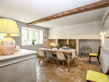 A dining room with a wooden table and six chairs near a window and a large fireplace at The Old Post Office in Coln St Aldwyns