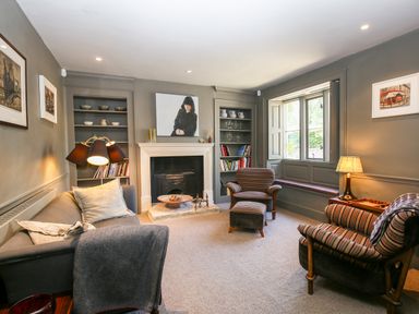 A living room with a fireplace two striped armchairs a gray sofa bookshelves and a window seat at The Old Post Office in Coln St Aldwyns