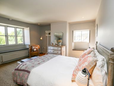 A bedroom with a bed pillows a dresser a chair a floor lamp and windows at The Old Post Office in Coln St Aldwyns
