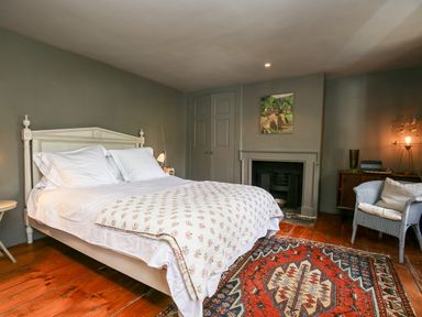 A bedroom with a white bed and pillows a patterned rug a fireplace a wicker chair and a wooden floor at The Old Post Office in Coln St Aldwyns