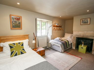A bedroom with two beds a wooden chest a floor lamp a fireplace and framed pictures at The Old Post Office in Coln St Aldwyns
