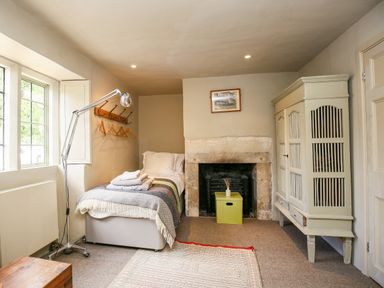 A bedroom with a single bed next to a window and a floor lamp a vintage wardrobe a fireplace with a green box inside and a painting above at The Old Post Office in Coln St Aldwyns